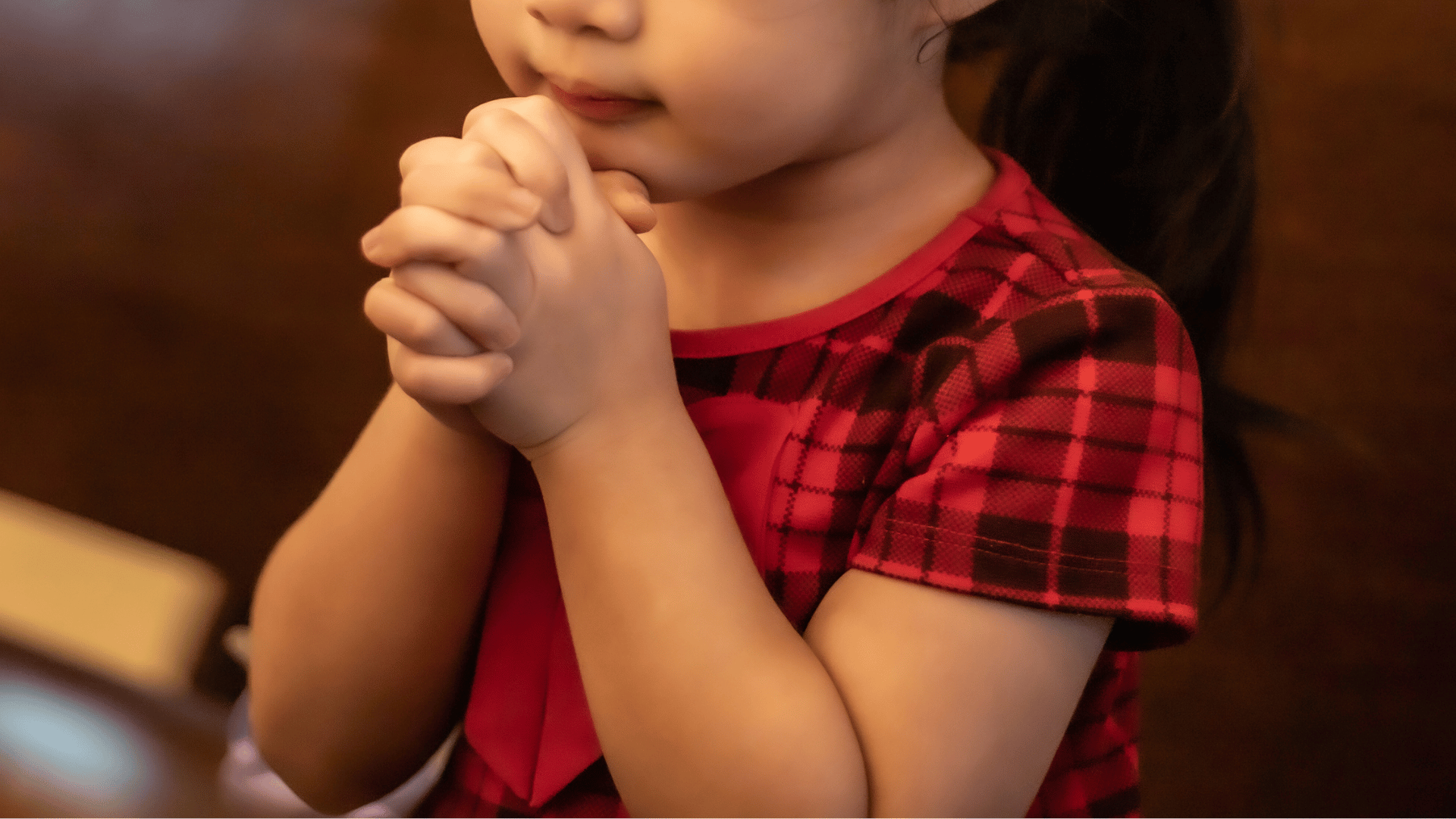 Criança rezando A imagem ilustra, em plano detalhe, uma criança sentada no banco de uma igreja rezando com as mãos juntas, próximas ao rosto.