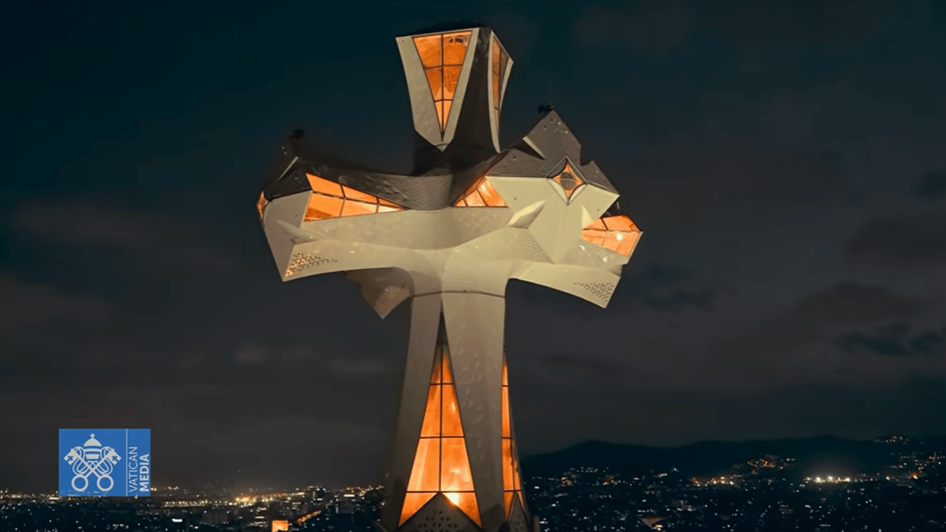 A imagem mostra, em ângulo fechado, bem de perto, a cruz iluminada no alto da torre central da basílica da sagrada familia