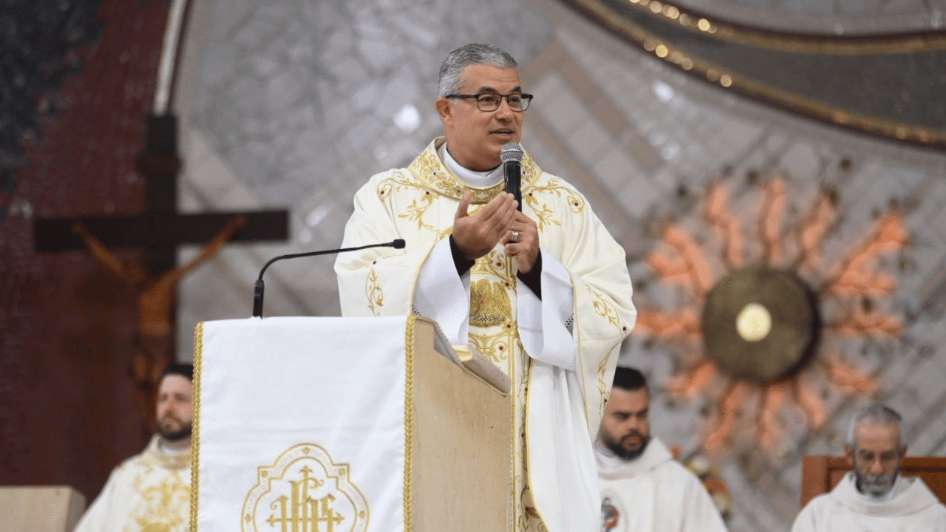 A imagem ilustra um padre católico segurando um microfone e discursando diante de um ambão, proferindo sua homilia. Ao fundo, vê-se um crucifixo, um sacrário e outros sacerdotes.