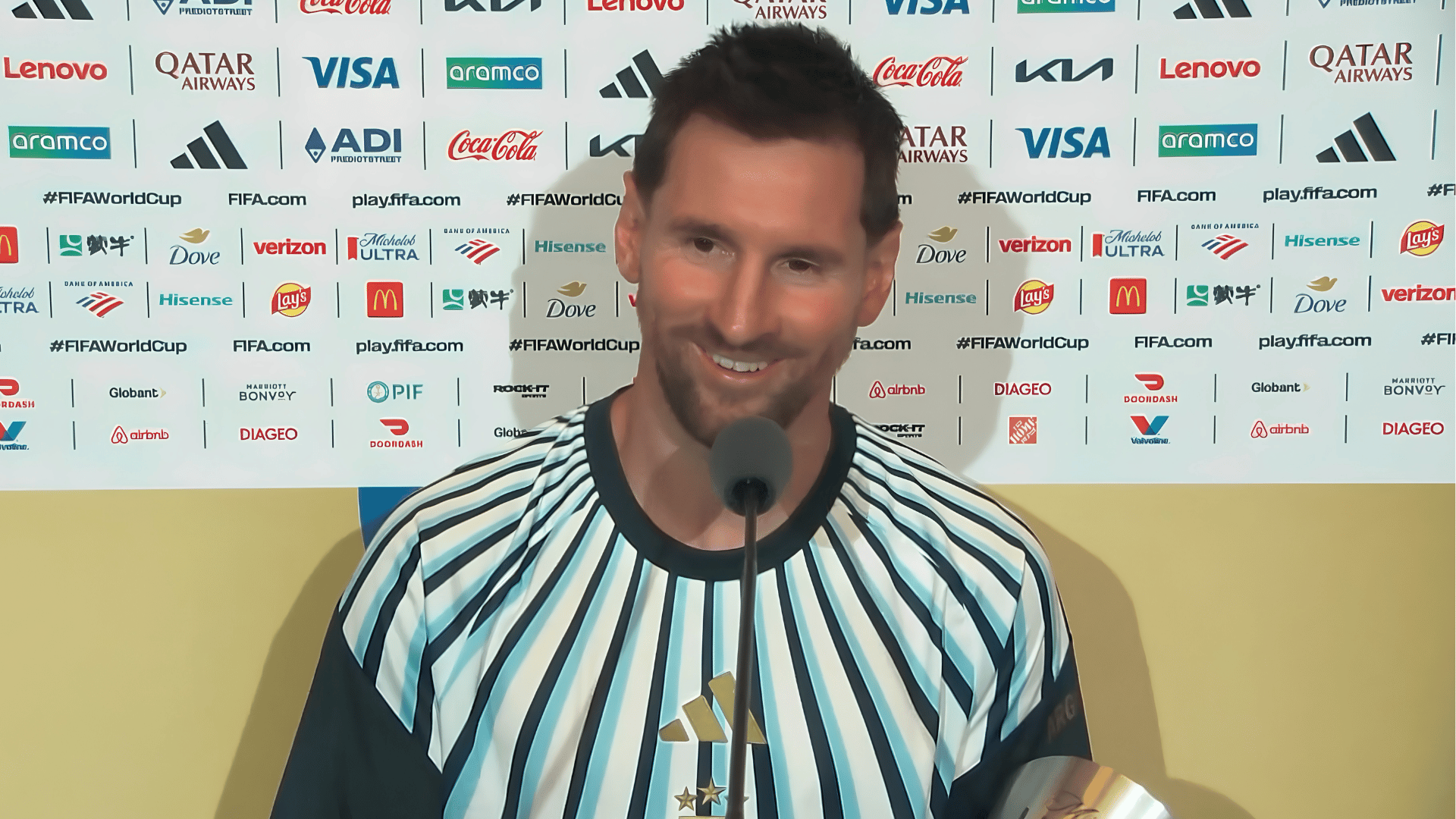 A imagem ilustra o atacante argentino Lionel Messi, vestido com uma camisa com listrar brancas e azuis, durante uma coletiva de imprensa.