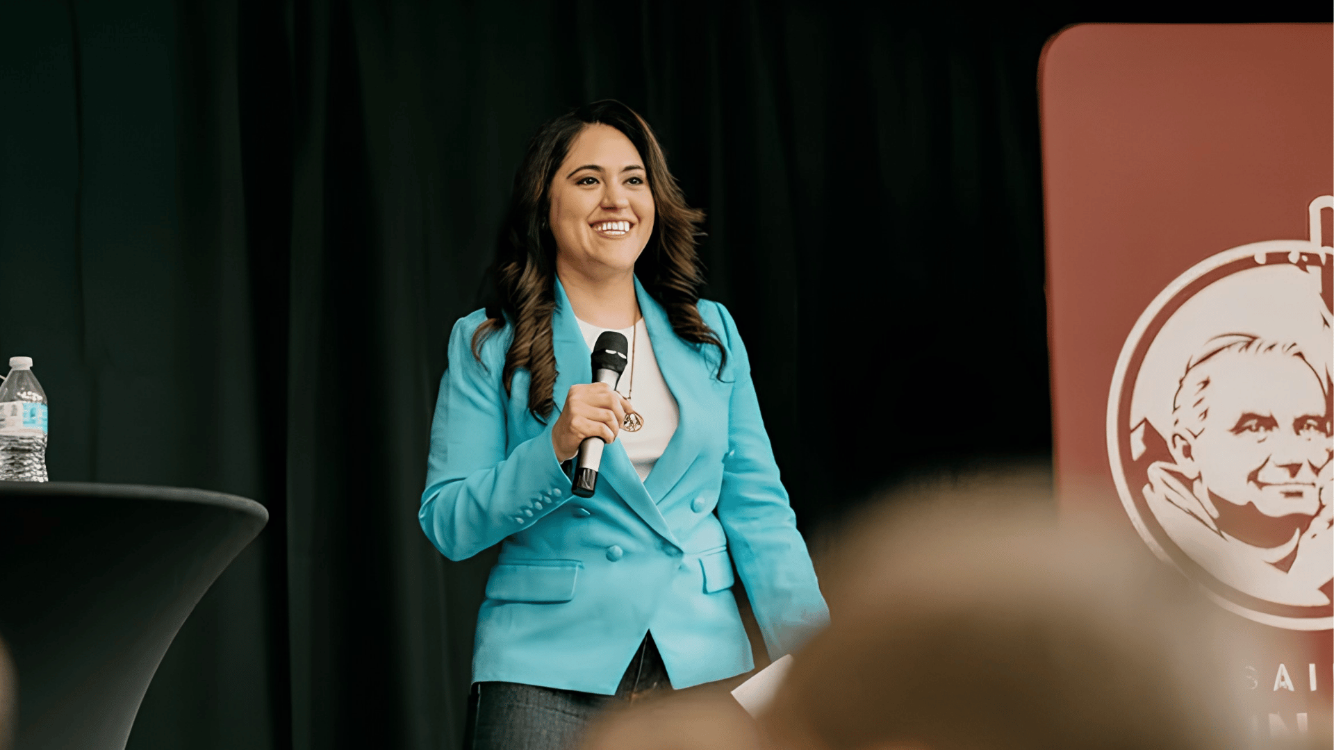 Maria Montserrat sorri enquanto fala ao público segurando um microfone durante um evento. Ela veste blazer azul-claro e está em um palco, ao lado de um painel com a imagem de São João Paulo II.