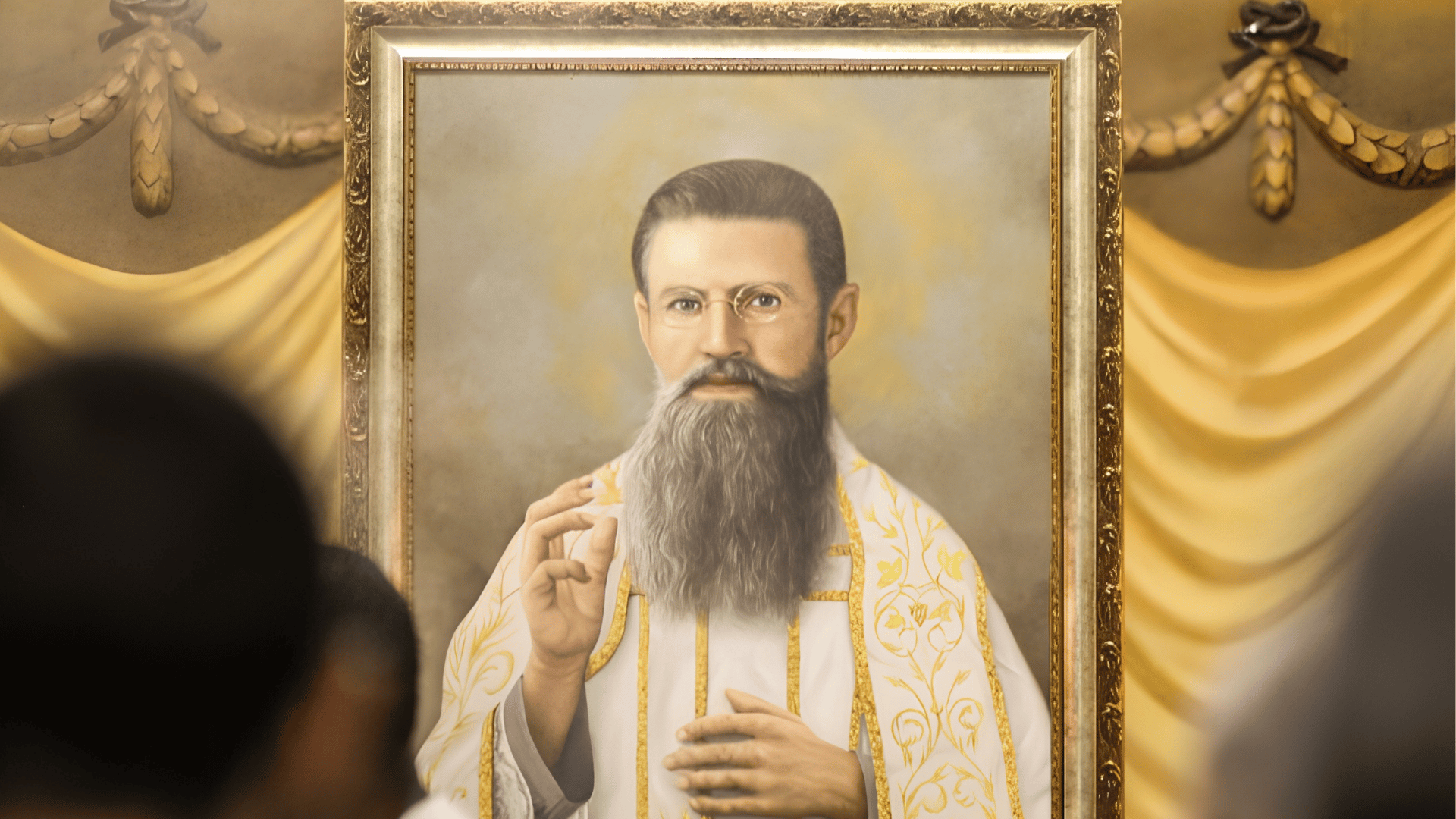 Retrato do padre Júlio Maria de Lombaerde emoldurado e exposto em ambiente religioso. O sacerdote aparece com vestes litúrgicas brancas ornamentadas em dourado, barba longa e óculos, fazendo um gesto de bênção. Ao fundo, há tecidos decorativos em tons dourados, enquanto fiéis aparecem desfocados em primeiro plano.