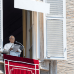 Papa Leão XIV faz a oração do Angelus da janela do Palácio Apostólico, no Vaticano. O Pontífice segura o texto da reflexão diante do microfone, enquanto a janela está aberta e ornamentada com uma tapeçaria vermelha.