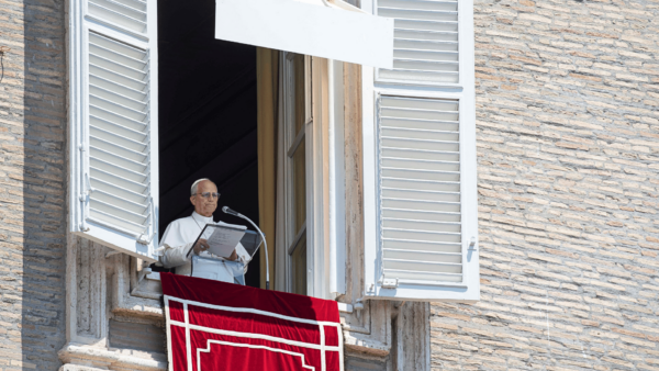 Papa Leão XIV faz a oração do Angelus da janela do Palácio Apostólico, no Vaticano. O Pontífice segura o texto da reflexão diante do microfone, enquanto a janela está aberta e ornamentada com uma tapeçaria vermelha.