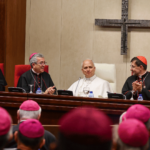 O Papa Leão XIV aparece com suas habituais vestes brancas. Ele está sentado a uma mesa com dois bispos ao seu lado e um crucifixo na parede ao fundo