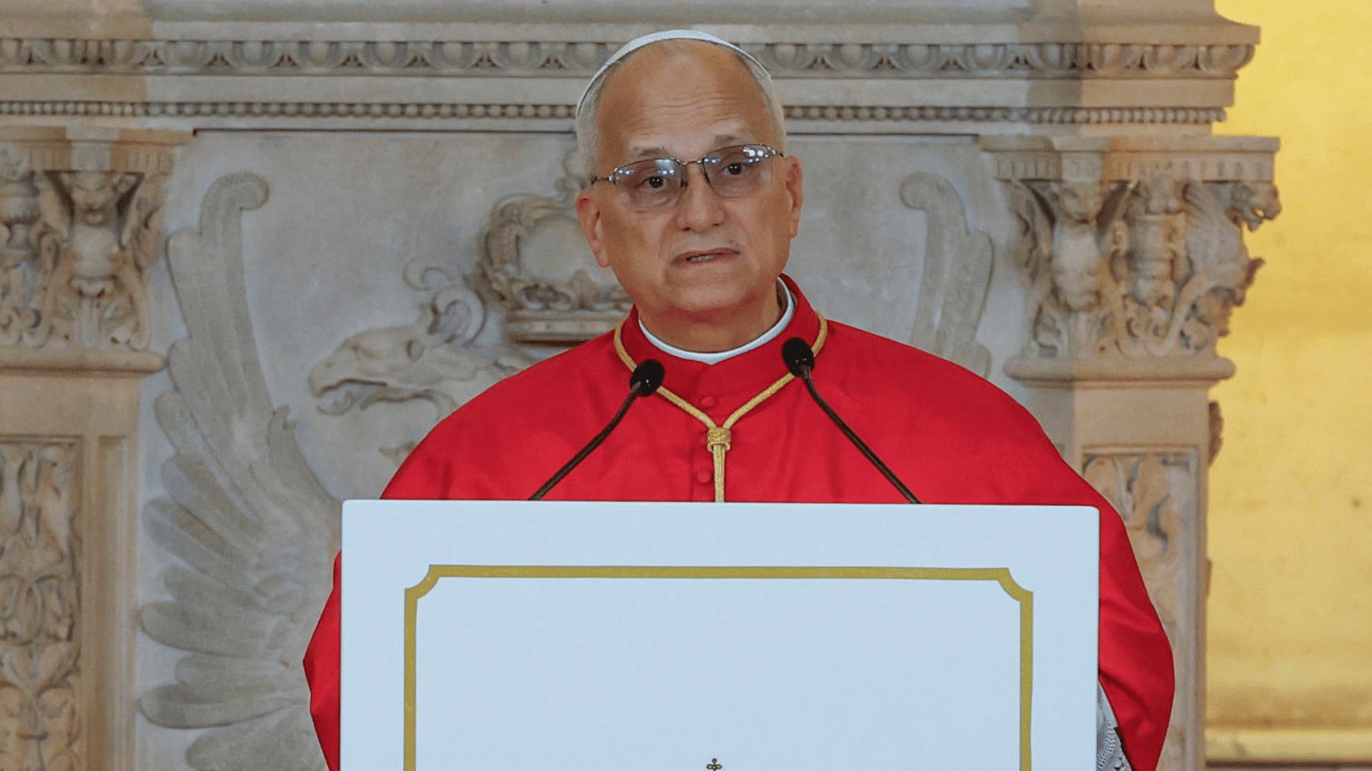 A imagem mostra o Papa Leão XIV com a moseta vermelha à frente de um púlpito com dois microfones