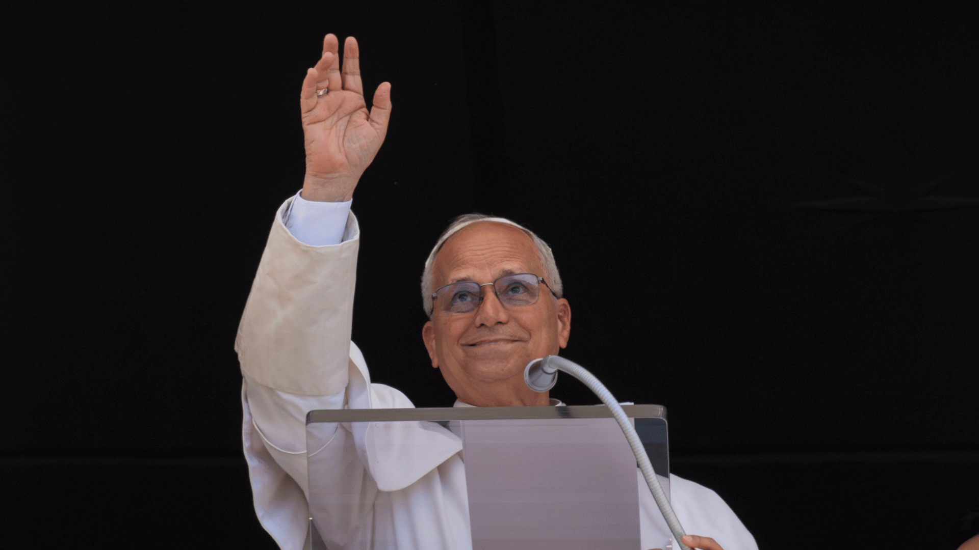 Papa Leão acena para os fiéis reunidos na Praça São Pedro A imagem ilustra o Papa Leão XIV, vestido com suas habituais vestes pontifícias, com o braço direito erguido em gesto de aceno. Ele está diante de um ambão e um microfone.