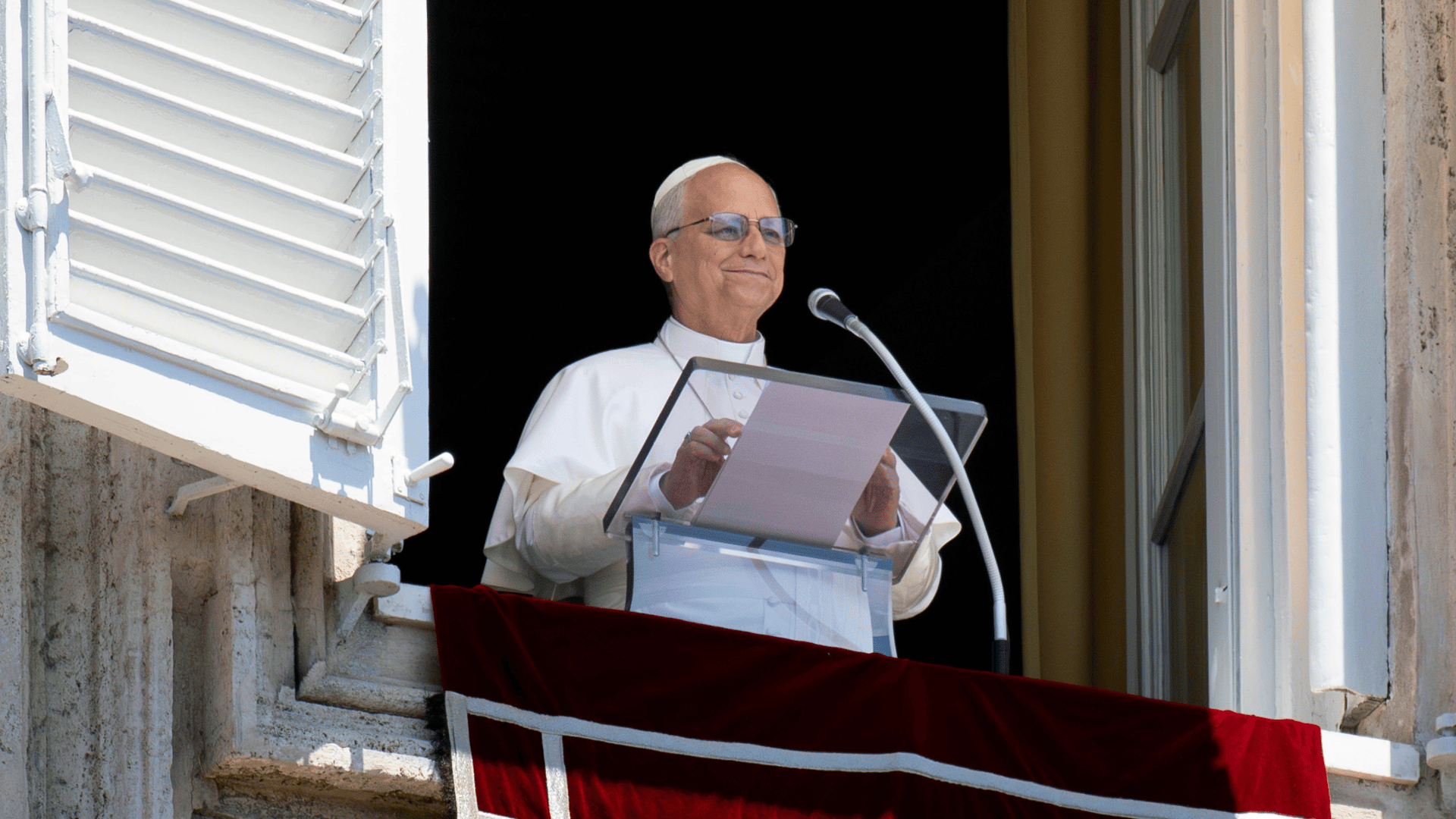 A imagem ilustra o Papa Leão XIV, com suas habituais vestes pontifícias, diante do parapeito de uma das janelas do Palácio Apostólico Vaticano, com um púlpito e um microfone à sua frente, durante a oração do Angelus.