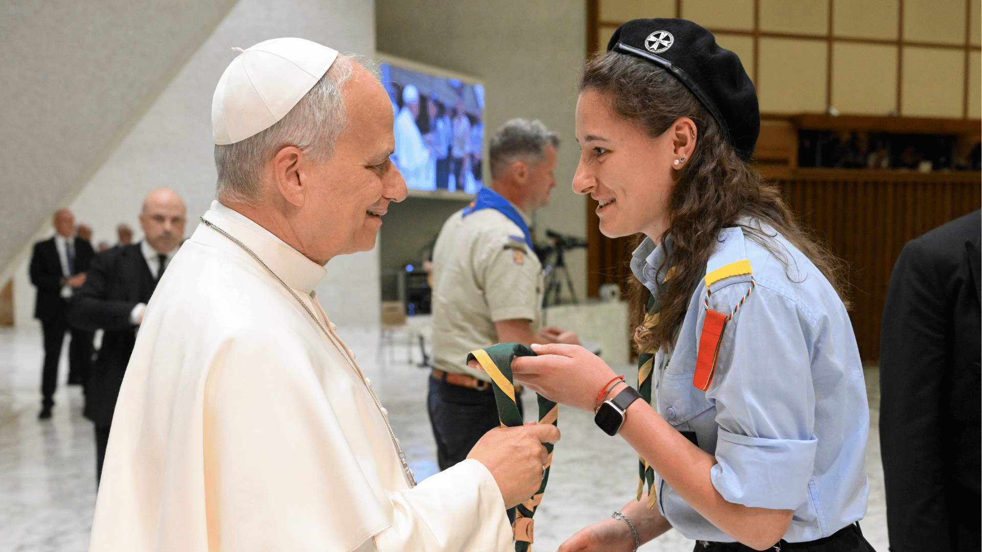 Papa Leão XIV conversa com uma jovem escoteira durante encontro no Vaticano. Os dois aparecem sorrindo e interagindo de forma próxima, enquanto outros participantes e membros da segurança estão ao fundo.