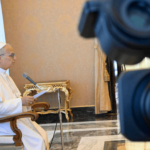 Papa Leão XIV em audiência no Vaticano. Sentado e com documentos em mãos, o Pontífice fala ao microfone enquanto câmeras registram a audiência.