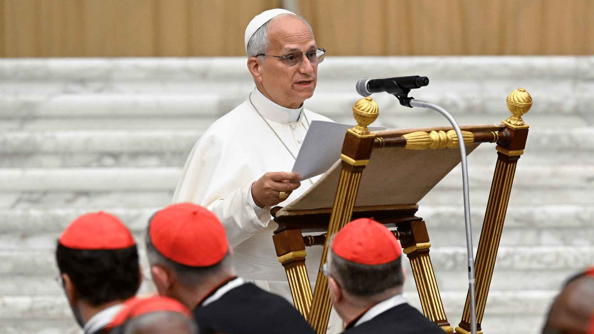 A imagem ilustra o Papa Leão XIV, com suas habituais vestes pontifícias, discursando diante de um púlpito com microfone. Alguns cardeais acompanham suas palavras.