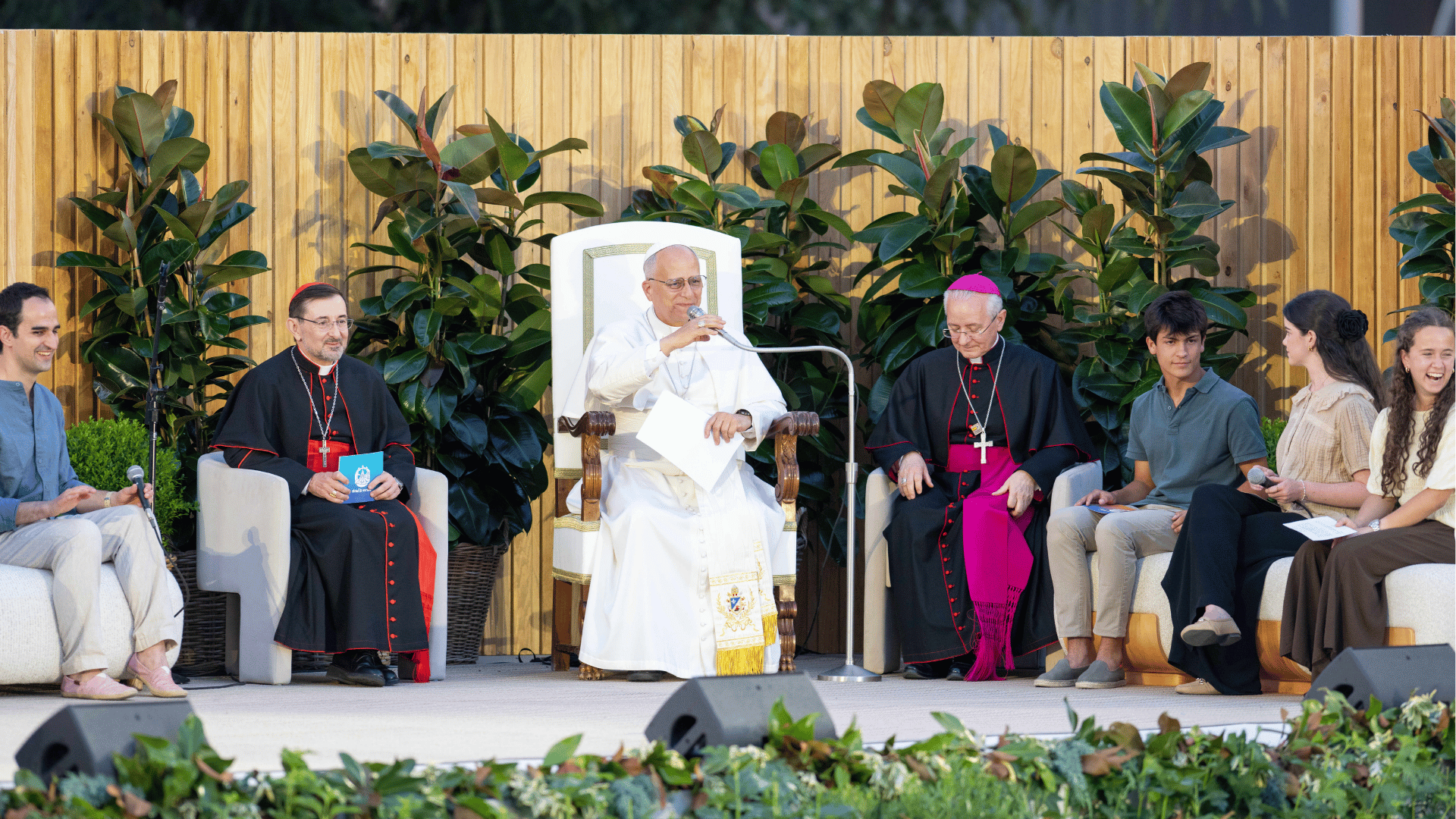 O Papa Leão XIV participa de um encontro com jovens em um palco ao ar livre. Sentado ao centro, ele fala ao microfone enquanto é acompanhado por cardeais, bispos e jovens que participam da conversa. Ao fundo, há uma estrutura de madeira decorada com plantas, criando um ambiente acolhedor para o diálogo.