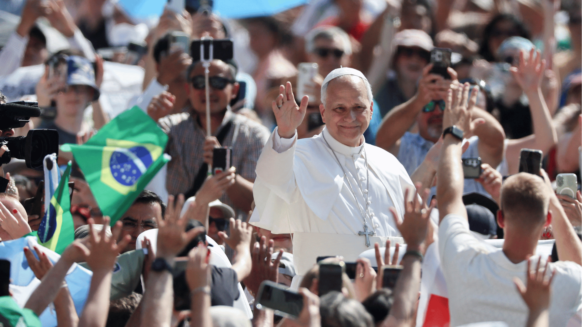Papa Leão XIV acena e abençoa os fiéis durante uma audiência pública ao ar livre. Cercado por uma multidão, o Pontífice aparece em veículo aberto enquanto peregrinos registram o momento com celulares e exibem bandeiras, entre elas a do Brasil.