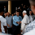 Papa Leão XIV cumprimenta um homem durante visita a uma unidade prisional na Espanha. Cercado por detentos, funcionários e profissionais da comunicação, o Pontífice estende a mão em gesto de proximidade e diálogo. A cena destaca o encontro pastoral com pessoas privadas de liberdade.