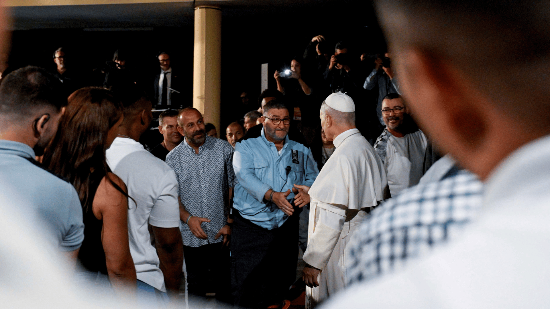 Papa Leão XIV cumprimenta um homem durante visita a uma unidade prisional na Espanha. Cercado por detentos, funcionários e profissionais da comunicação, o Pontífice estende a mão em gesto de proximidade e diálogo. A cena destaca o encontro pastoral com pessoas privadas de liberdade.