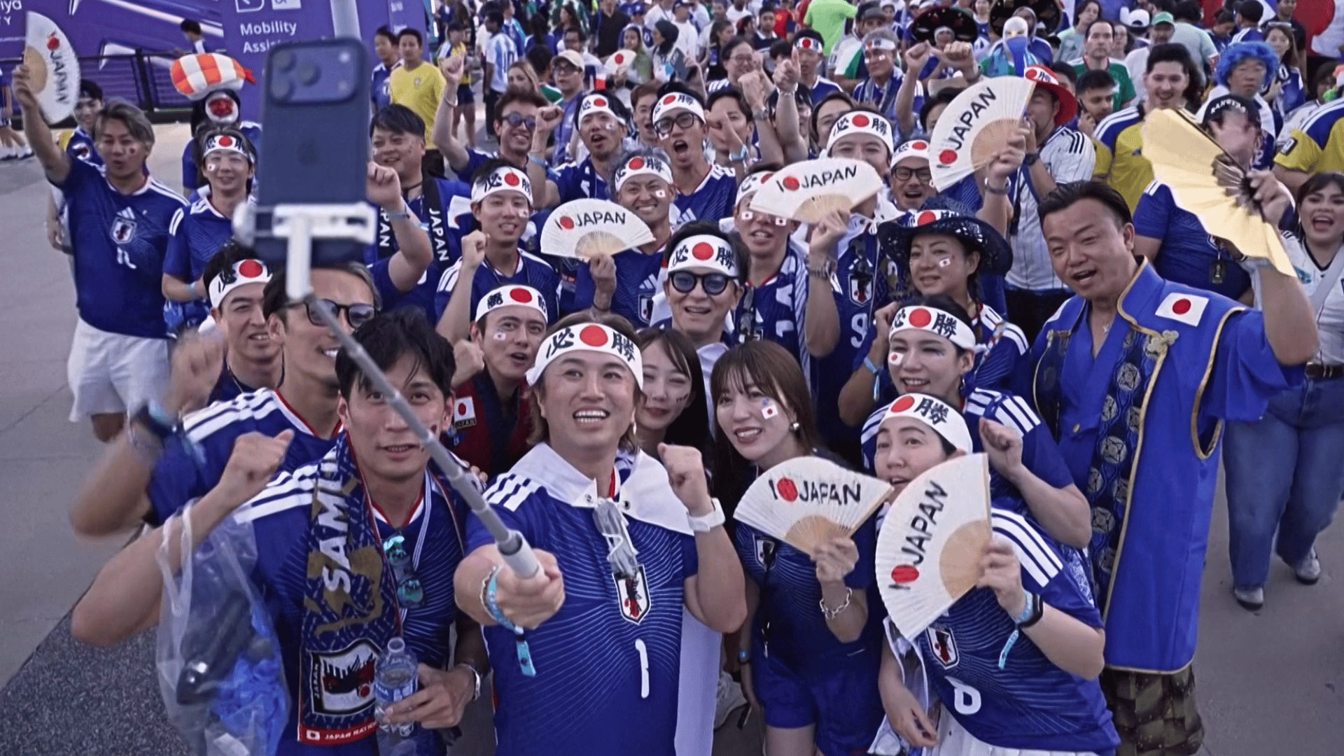 A imagem ilustra torcedores do Japão tirando uma selfie do lado de fora de um estádio.