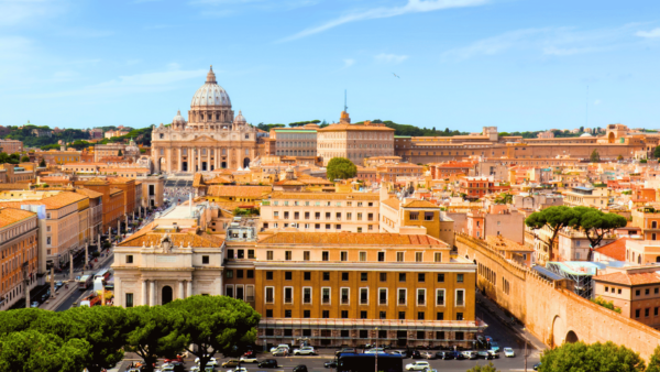 Vista panorâmica da Cidade do Vaticano, com destaque para a Basílica de São Pedro ao centro, cercada por edifícios históricos, muralhas do Vaticano e árvores sob céu azul.