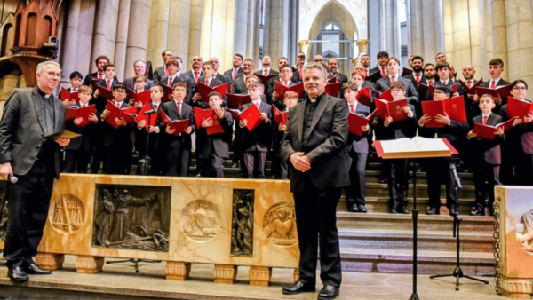 Coral da Capela Sistina reunido no altar da Catedral da Sé, em São Paulo, durante apresentação da turnê no Brasil. À frente do grupo estão dois sacerdotes, enquanto os coralistas, entre crianças, adolescentes e adultos, seguram partituras vermelhas.