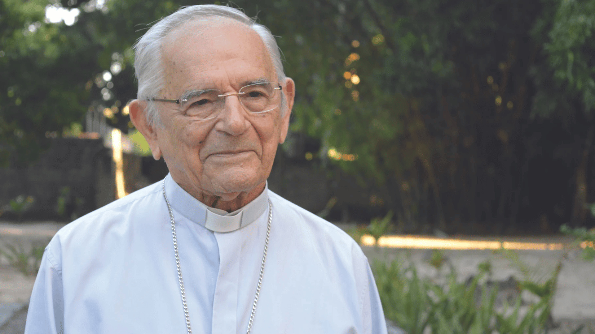 Dom Heitor usa uma camisa de manga longa azul claro com colarinho clerical branco (clérgima) e óculos. Ao seu fundo há uma vegetação