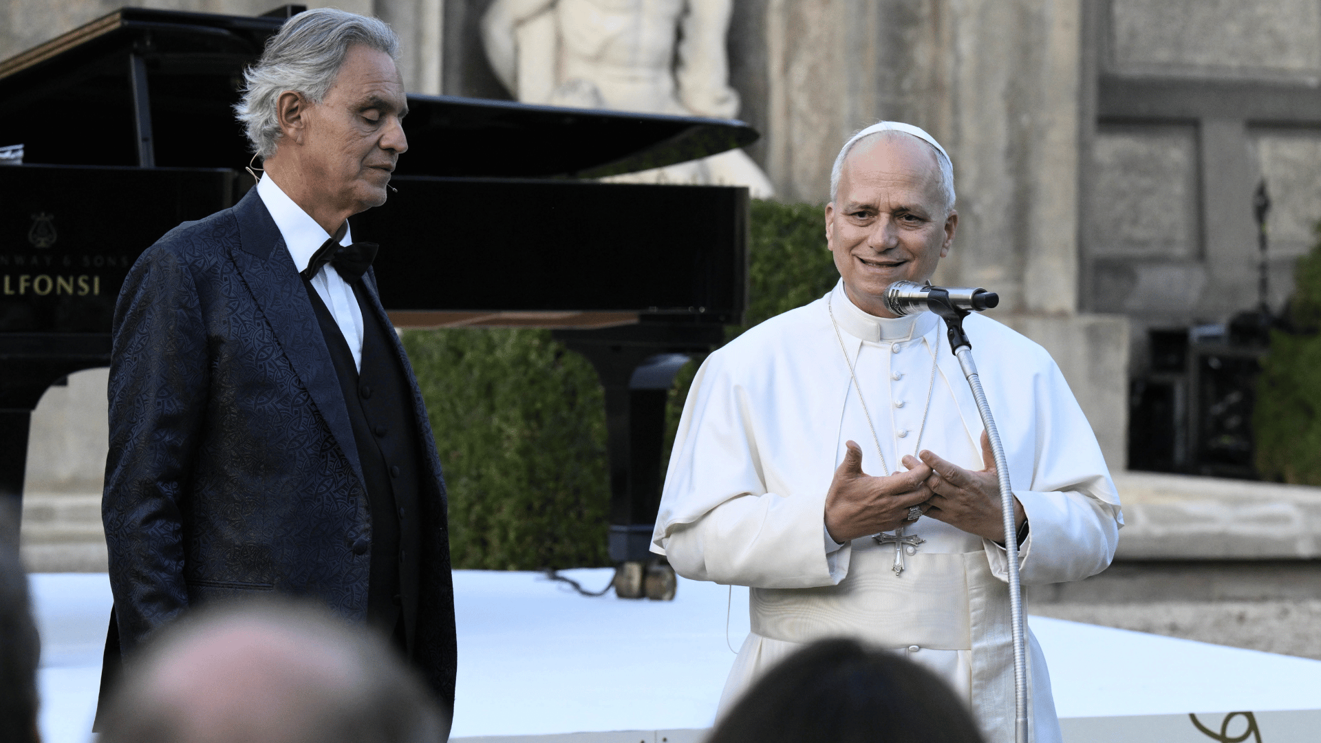 O Papa Leão XIV aparece com suas habituais vestes brancas em pé diante de um microfone. Ao seu lado, está o tenor Andrea Bocceli com smoke preto. 