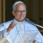 O Papa Leão XIV aparece com suas habituais vestes brancas diante de um púlpito com microfone. Ele acena com a mão direita