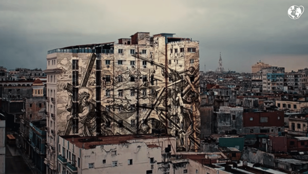 Vista panorâmica de uma cidade e um grande mural, com uma imagem sobreposta na fachada de um edifício, retratando Jesus carregando a cruz e outras cenas da Paixão de Cristo.
