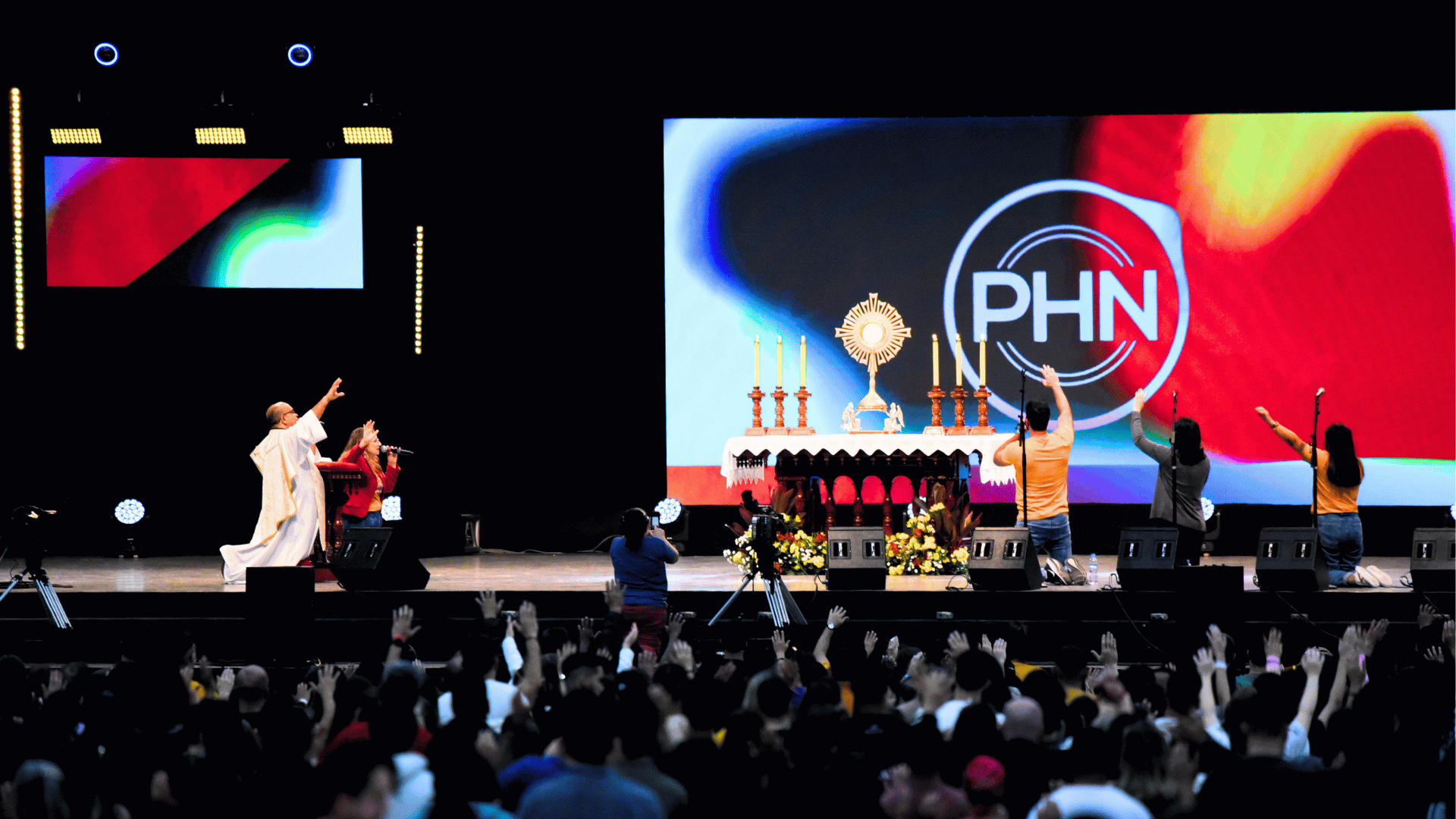 Momento de adoração ao Santíssimo Sacramento durante o PHN 2024, com fiéis de braços erguidos diante do altar no palco principal do evento.
