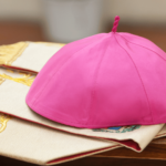Solidéu roxo de bispo sobre vestes litúrgicas brancas com detalhes dourados, símbolo do ministério episcopal na Igreja Católica.