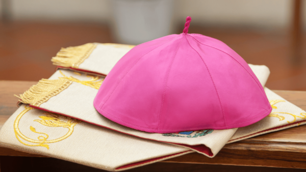 Solidéu roxo de bispo sobre vestes litúrgicas brancas com detalhes dourados, símbolo do ministério episcopal na Igreja Católica.