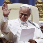 A imagem mostra o Papa Leão XIV com suas habituais vestes brancas aparece sentado na tradicional cadeira pontifícia usada para a catequese. Ele segura um papel em uma das mãos e com a outra acena para os fiéis