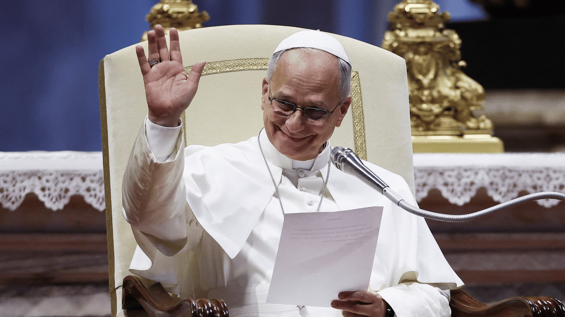 A imagem mostra o Papa Leão XIV com suas habituais vestes brancas aparece sentado na tradicional cadeira pontifícia usada para a catequese. Ele segura um papel em uma das mãos e com a outra acena para os fiéis
