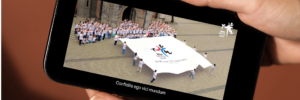 Pessoa segura um celular exibindo uma cena do videoclipe do hino oficial da Jornada Mundial da Juventude (JMJ) Seul 2027, com jovens reunidos em uma praça ao redor da bandeira do evento.