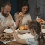 A foto mostra uma família reunida à mesa: há um homem, uma mulher e duas crianças (um menino e uma menina). Todos estão de mãos dadas, como se estivesse rezando antes da refeição. A mesa possui travessas com biscoitos, bolo e taças com sucos.