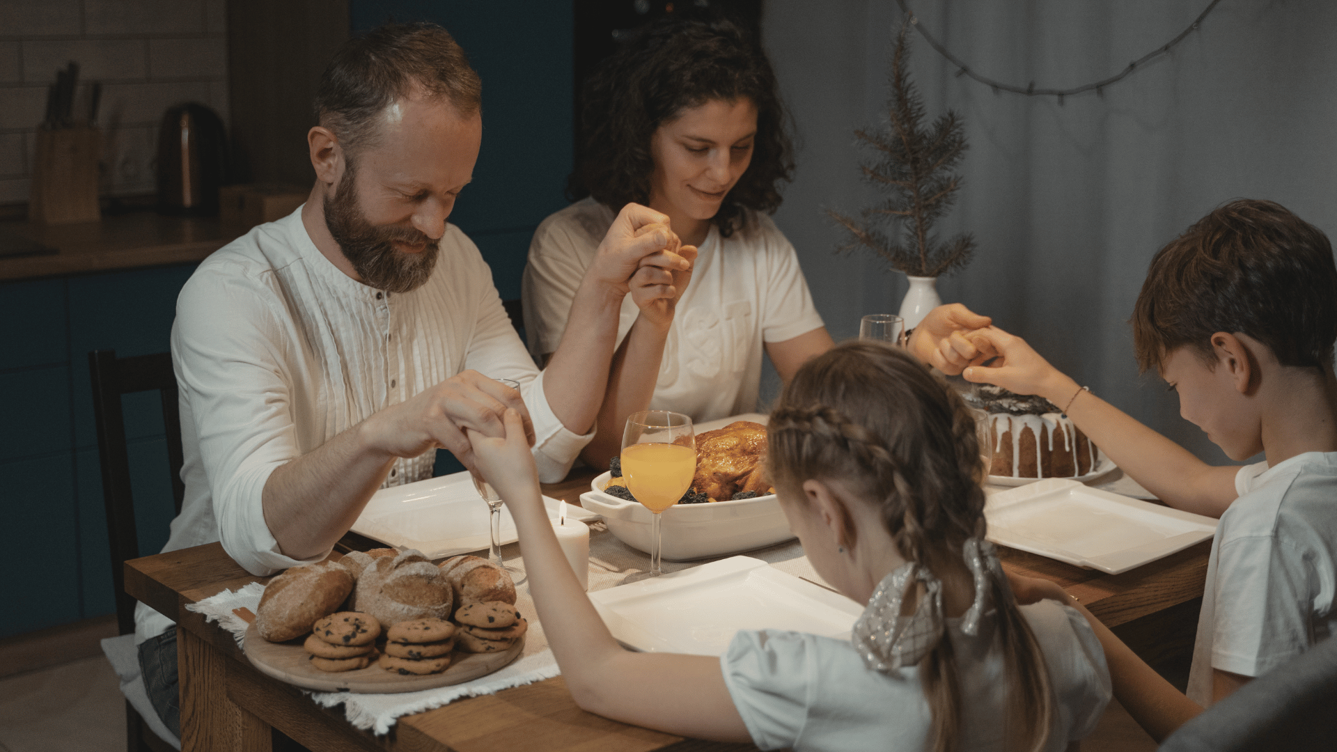 A foto mostra uma família reunida à mesa: há um homem, uma mulher e duas crianças (um menino e uma menina). Todos estão de mãos dadas, como se estivesse rezando antes da refeição. A mesa possui travessas com biscoitos, bolo e taças com sucos.