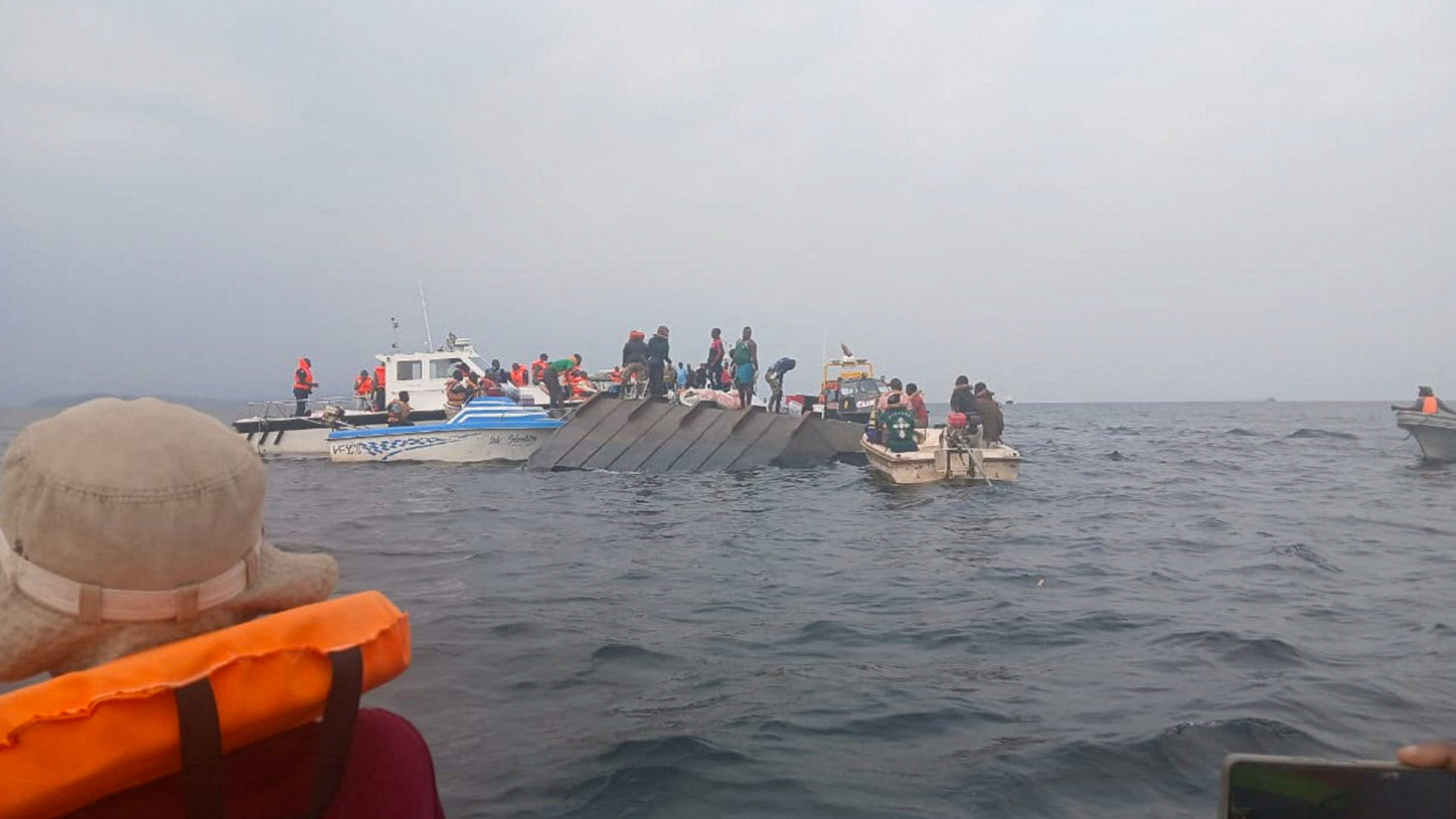 A imagem mostra uma embarcação virada no mar e outra embarcação da equipe de resgate que trabalham com coletes laranjas