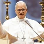 A imagem mostra o Papa Leão XIV com suas habituais vestes brancas, de mãos elevadas, braços abertos, diante de um microfone prateado