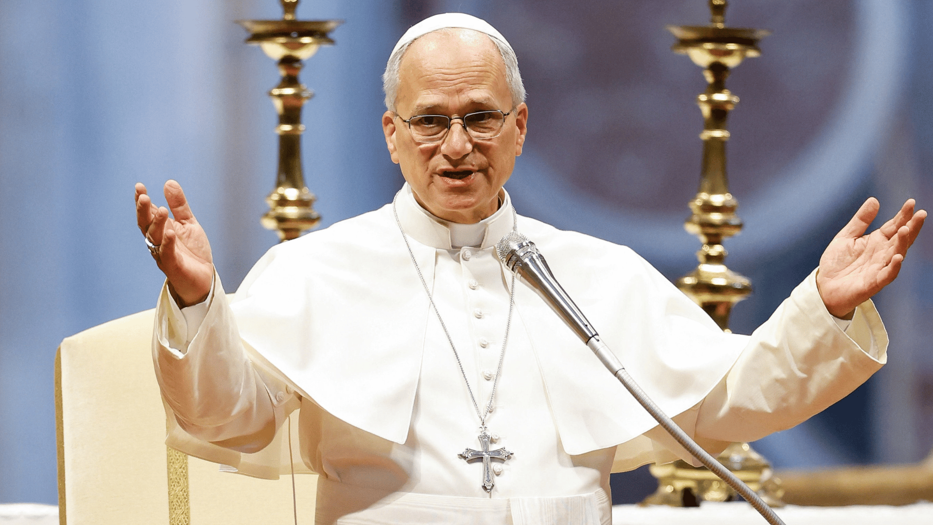 A imagem mostra o Papa Leão XIV com suas habituais vestes brancas, de mãos elevadas, braços abertos, diante de um microfone prateado