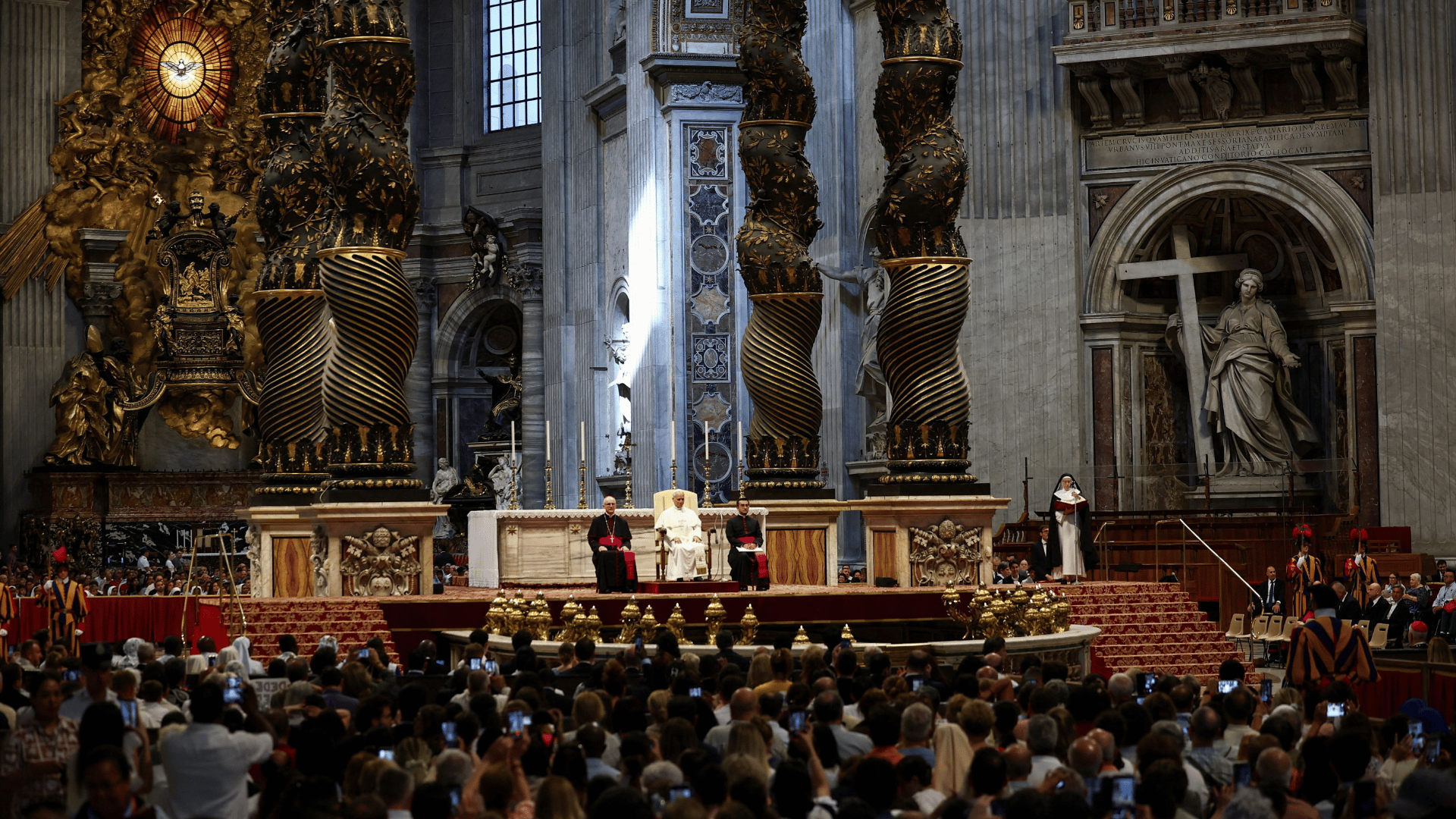 A imagem mostra, em plano aberto, a Basílica de São Pedro. No altar, está o Papa sentado e uma cadeira e dois clérigos sentados ao seu lado. Diante deles, estão os fiéis.
