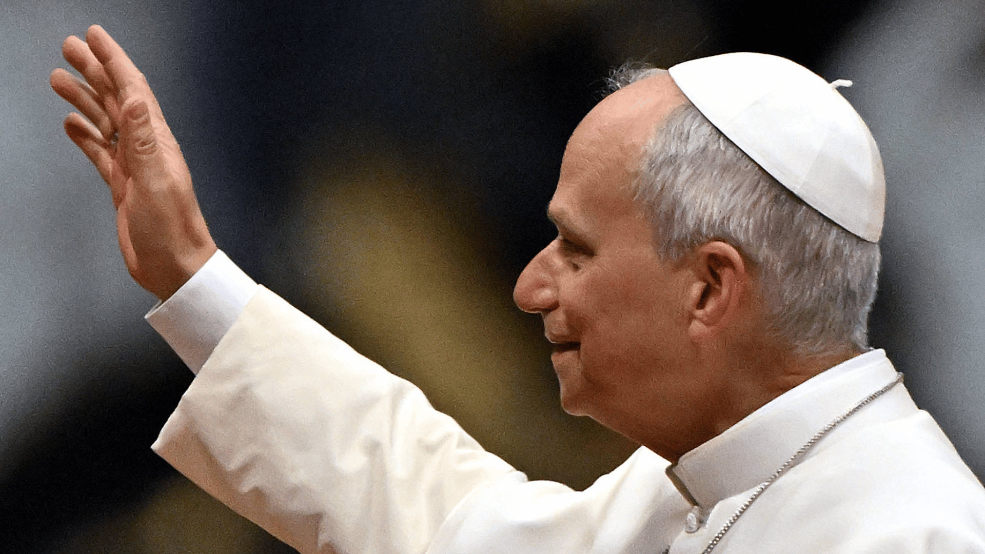 A imagem ilustra o Papa Leão XIV, de perfil, com suas habituais vestes pontifícias, com o braço direito erguido em um gesto de aceno.