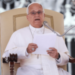 A foto mostra o Papa Leão XIV com suas habituais vestes brancas sentado na tradicional cadeira utilizada para a Audiência Geral (uma cadeira bege com braços de madeira). O Papa está sentado diante de um microfone e segura um papel nas mãos.