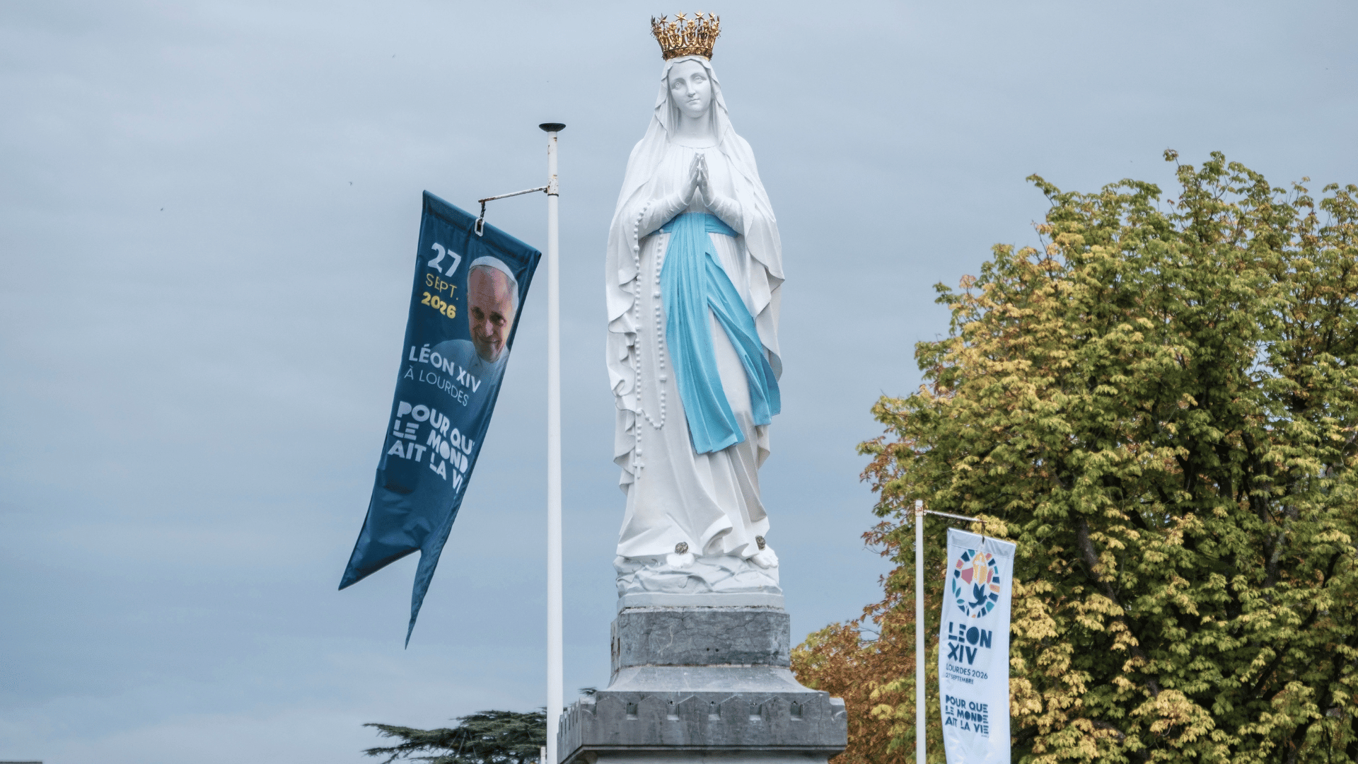 A imagem mostra uma imagem de Nossa Senhora de Lourdes ao lado da qual aparecem dois banners de divulgação sobre a visita do Papa Leão XIV