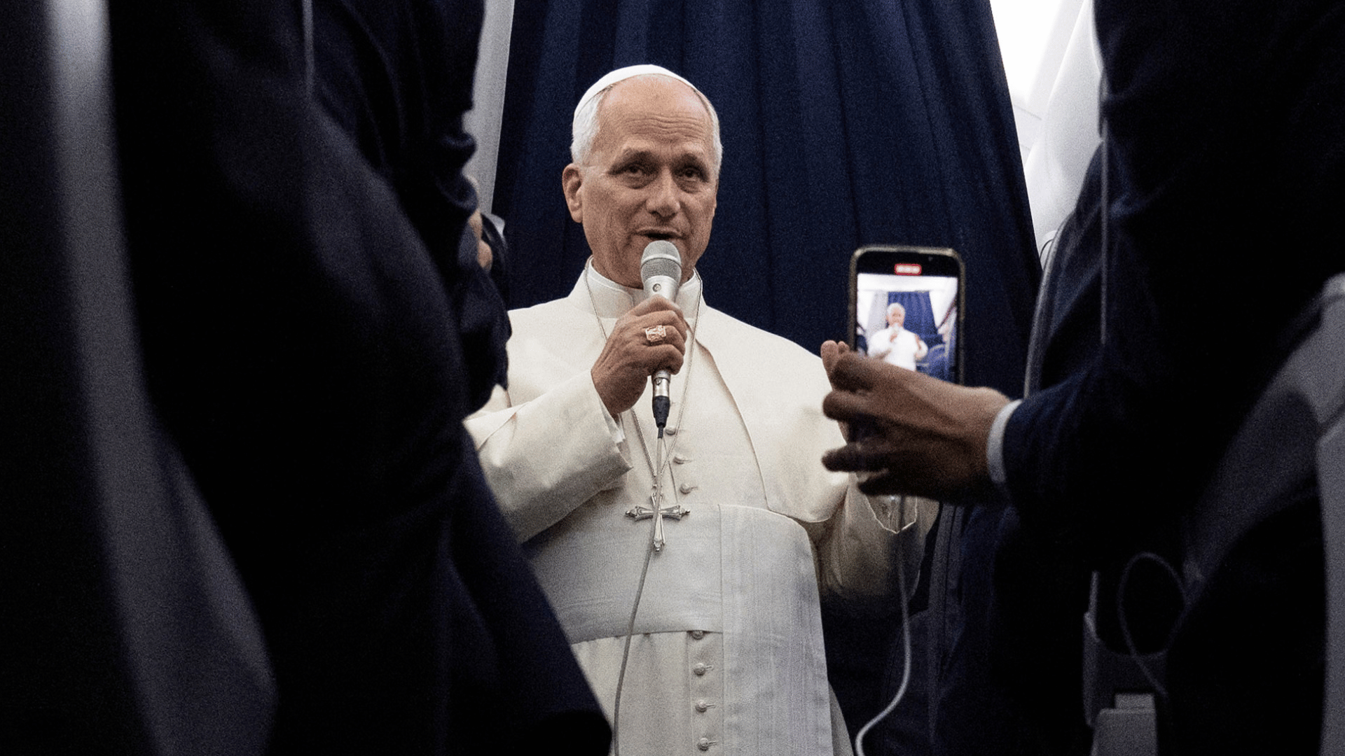 A imagem mostra o Papa Leão XIV em pé dentro do avião. Ele segura um microfone e fala aos jornalistas que o acompanham no voo.