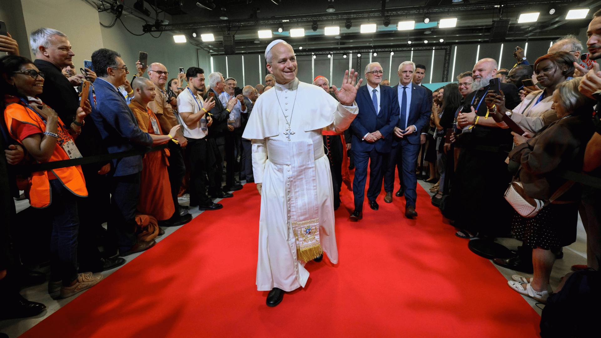 A imagem mostra o Papa Leão XIV entrando em uma sala cheia de pessoas. Ele está com suas habituais vestes brancas e acena para as pessoas sorridente, caminhando sobre um tapete vermelho.
