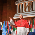 Papa acena durante visita à Unesco, em Paris. Atrás dele estão as bandeiras dos países que integram a organização, além do símbolo da Unesco.
