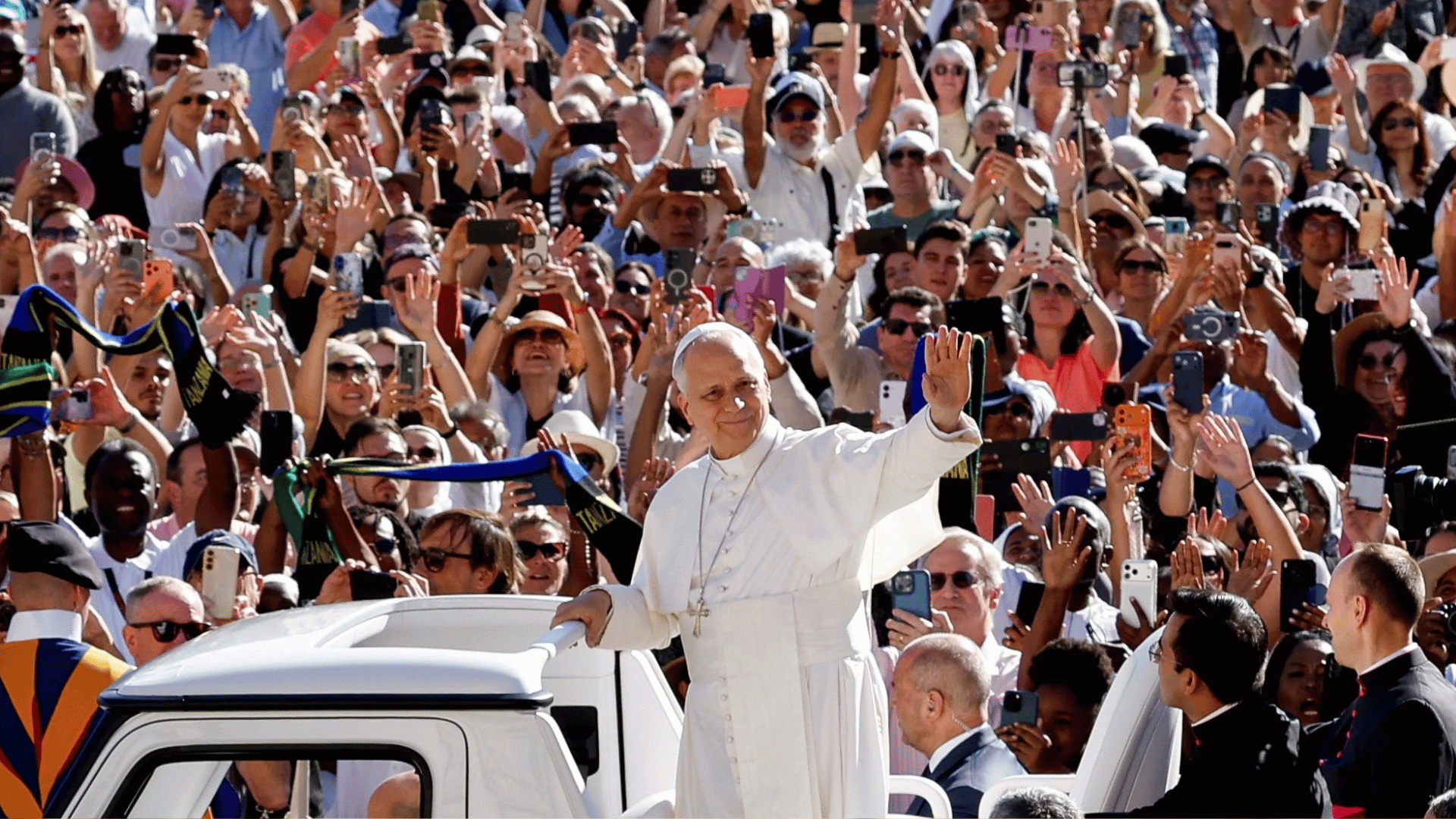 A imagem mostra o Papa com suas habituais vestes brancas em pé no papamóvel acenando para uma multidão de fiéis na Praça São Pedro