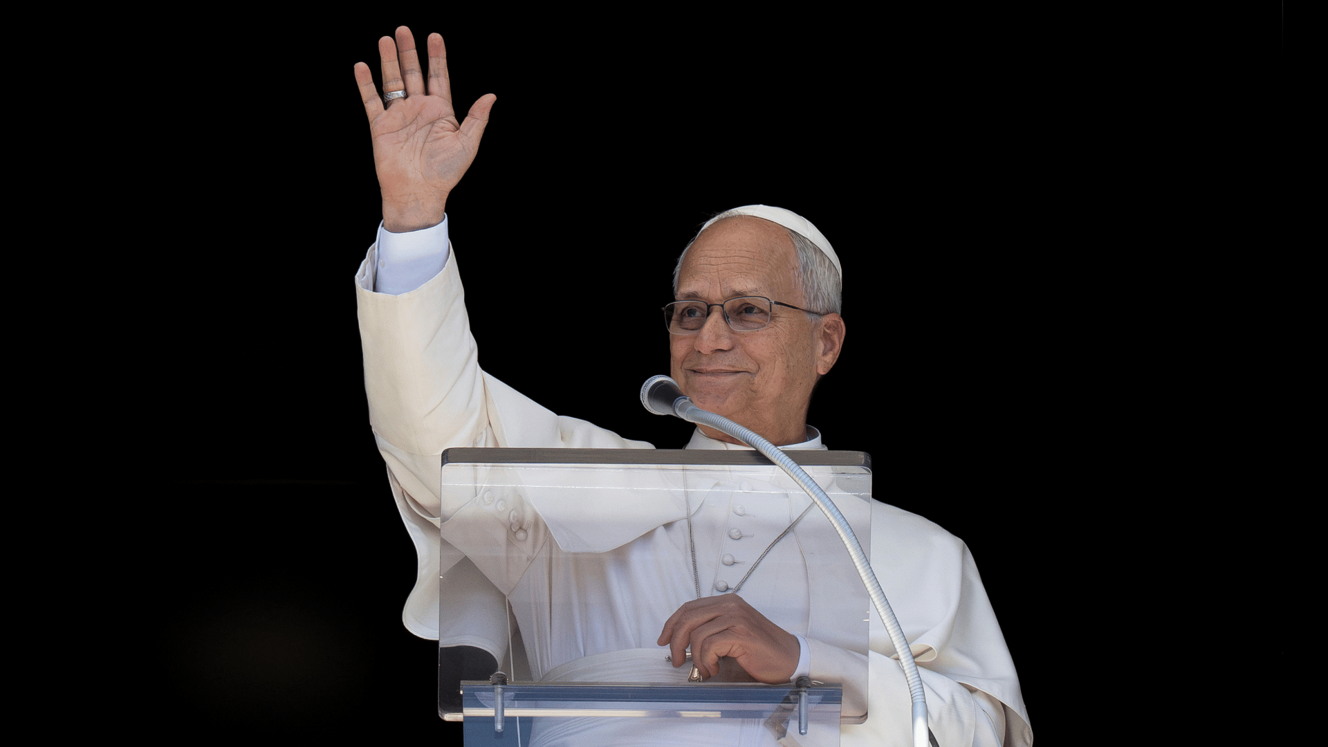 Papa Leão XIV no Angelus A imagem ilustra o Papa Leão XIV, com suas habituais vestes pontifícias, diante de um púlpito e um microfone. Ele tem o braço direito erguido, acenando.
