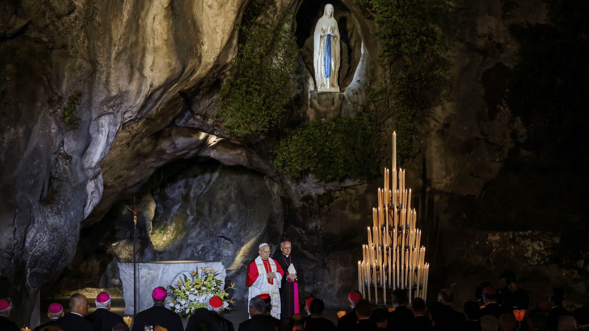A imagem mostra o Papa Leão XIV aos pés da imagem de Nossa Senhora na gruta de Lourdes, na França. Além dele, há membros do clero e fiéis segurando velas nas mãos