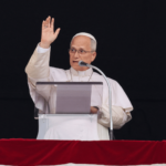 A imagem mostra o Papa Leão XIV na janela do palácio apostólico vaticano. Ele acena aos fiéis e fala ao microfone.