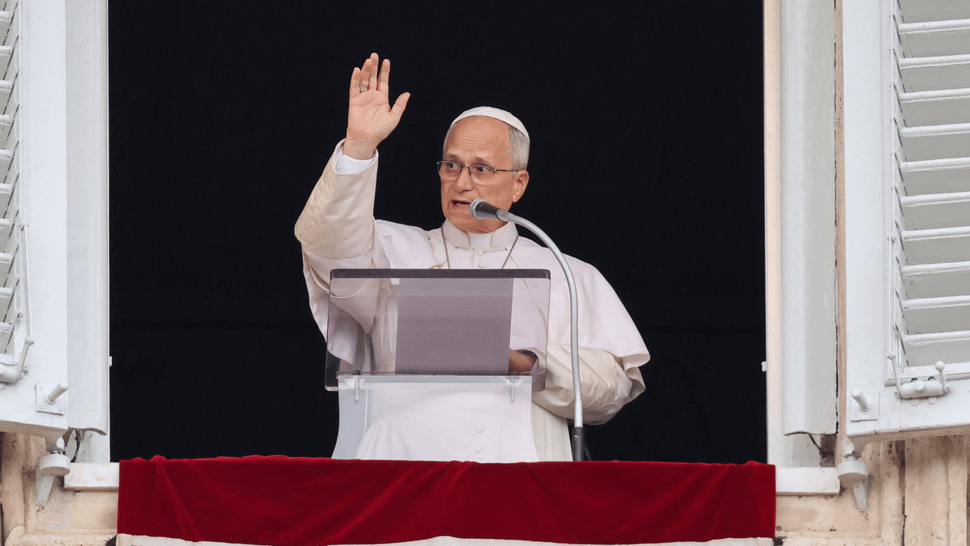 A imagem mostra o Papa Leão XIV na janela do palácio apostólico vaticano. Ele acena aos fiéis e fala ao microfone.