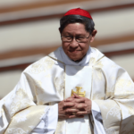 Cardeal Luis Antonio Tagle, com paramentos litúrgicos brancos e solidéu vermelho, mantém as mãos unidas durante celebração.